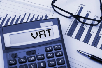 Calculator displaying "VAT" on screen with financial charts, glasses, and a pen in the background.